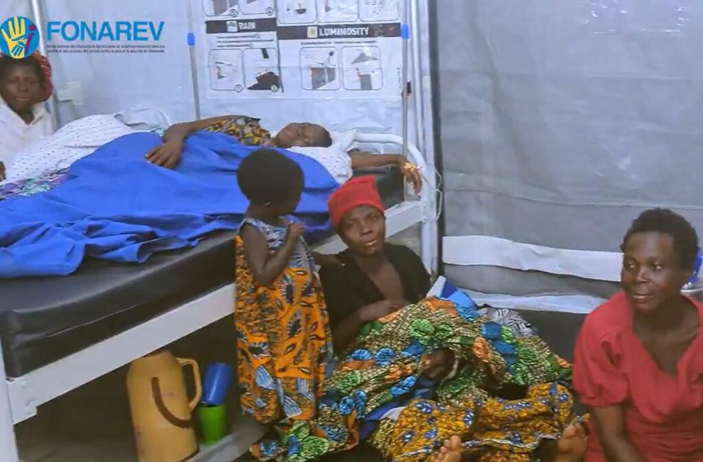 Mwanamke na mtoto wamekaa kando ya kitanda katika hema la matibabu huko Busuma, Burundi, wakati mtoto mwingine amelala chini ya blanketi ya bluu.