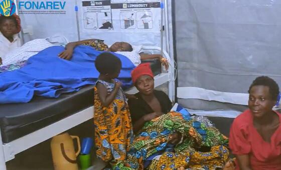 Mwanamke na mtoto wamekaa kando ya kitanda katika hema la matibabu huko Busuma, Burundi, wakati mtoto mwingine amelala chini ya blanketi ya bluu.