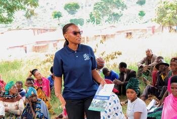 Mwanamke aliyevaa kanzu ya polo ya WCLAC na miwani ya jua anazungumza na watazamaji walioketi katika mazingira ya vijijini Tanzania, akibeba vifaa vya elimu.