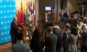 Secretary-General António Guterres speaks at a podium during a press conference about the US-Israel-Iran conflict, with multiple national flags and security council logos in the background.