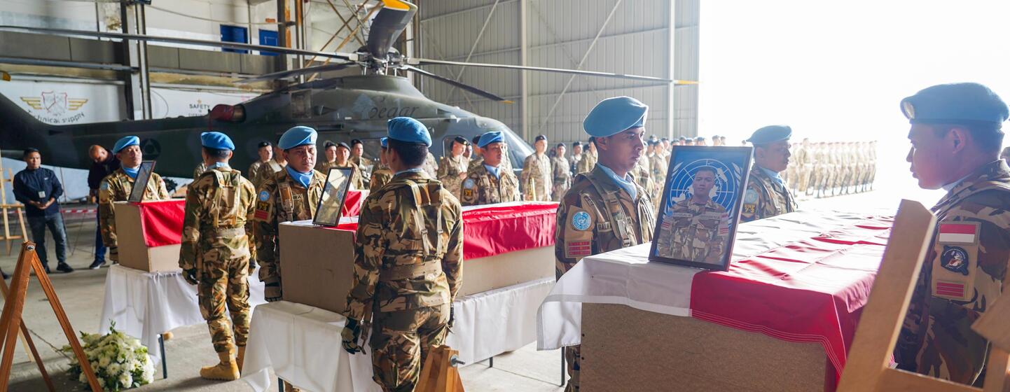 Des soldats des forces de maintien de la paix de l'UNIFIL en bérets bleus et uniformes de camouflage se tiennent en formation à côté de cercueils couverts lors d'une cérémonie funèbre dans un hangar avec un hélicoptère à l'arrière-plan.