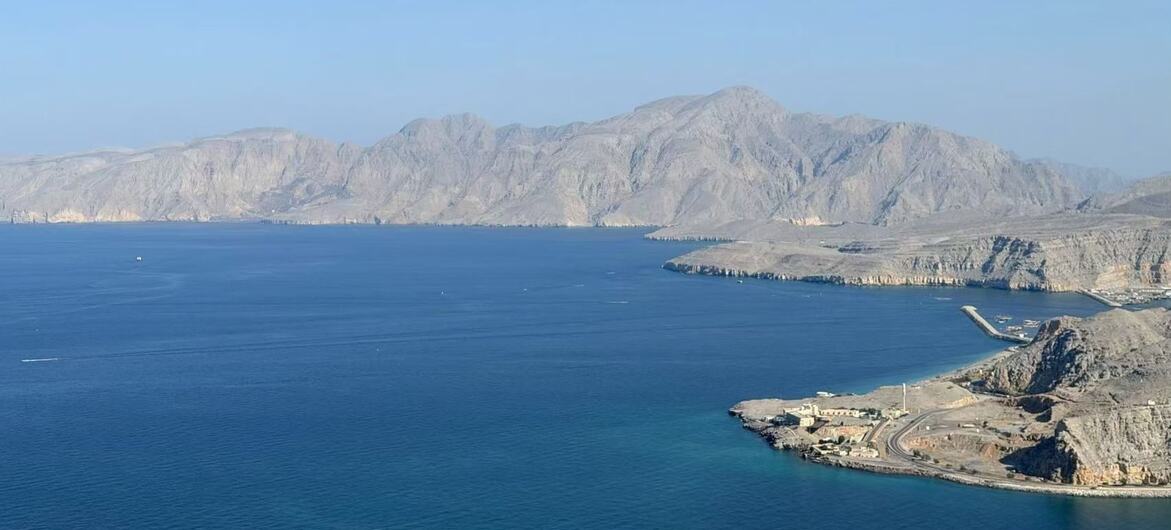 A wide bay with deep blue water, bordered by arid, rocky mountains and a small coastal settlement on the right.