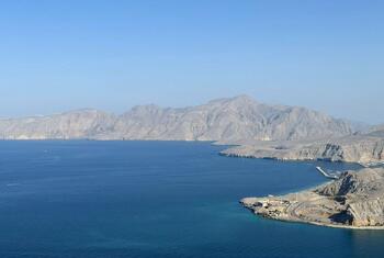 A wide bay with deep blue water, bordered by arid, rocky mountains and a small coastal settlement on the right.