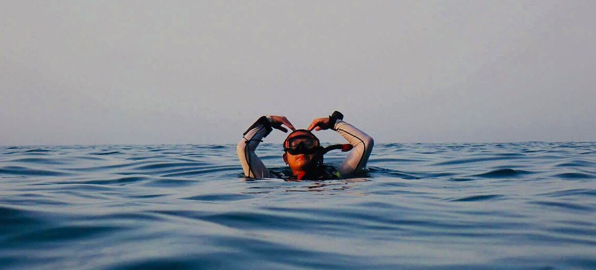 A person in a wetsuit and goggles floats in the ocean, adjusting their mask with both hands.