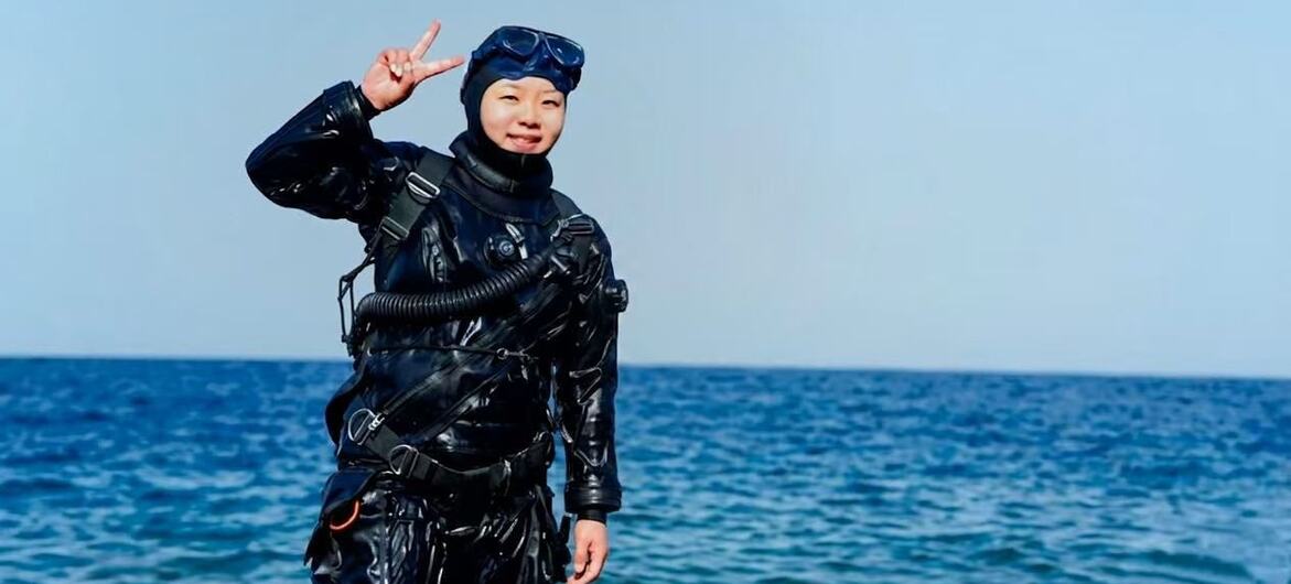 A person in a scuba diving suit and mask makes a peace sign against the ocean and blue sky.