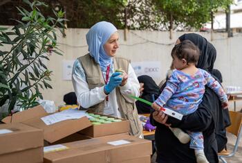 Mfanyakazi wa UNICEF aliyevalia hijab ya bluu nyepesi na koti, akiwa amevaa glavu za bluu, anapima ukuaji wa mtoto aliyebebwa na mama katika makao ya muda huko Beirut, Lebanoni.