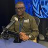 Mtu mwenye miwani ameketi kwenye dawati na kipaza sauti katika studio, akiwa na nembo ya Umoja wa Mataifa kwenye skrini nyuma yake.