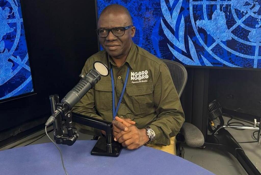 Mtu mwenye miwani ameketi kwenye dawati na kipaza sauti katika studio, akiwa na nembo ya Umoja wa Mataifa kwenye skrini nyuma yake.