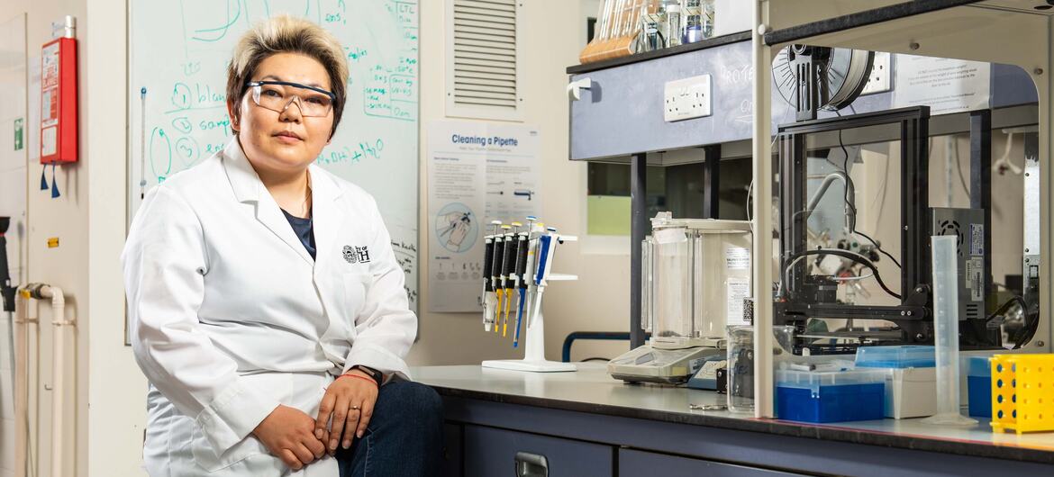 Ученый в белом халате сидит в лаборатории с научным оборудованием и доской на заднем плане.