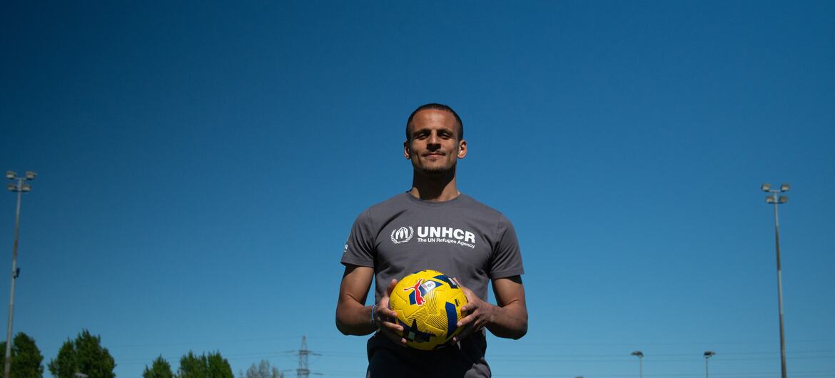 足球运动员阿里·哈马迪（Ali Al-Hamadi）身穿一件灰色的联合国难民署（UNHCR）T 恤，在阳光明媚的足球场上手持一个黄色足球。