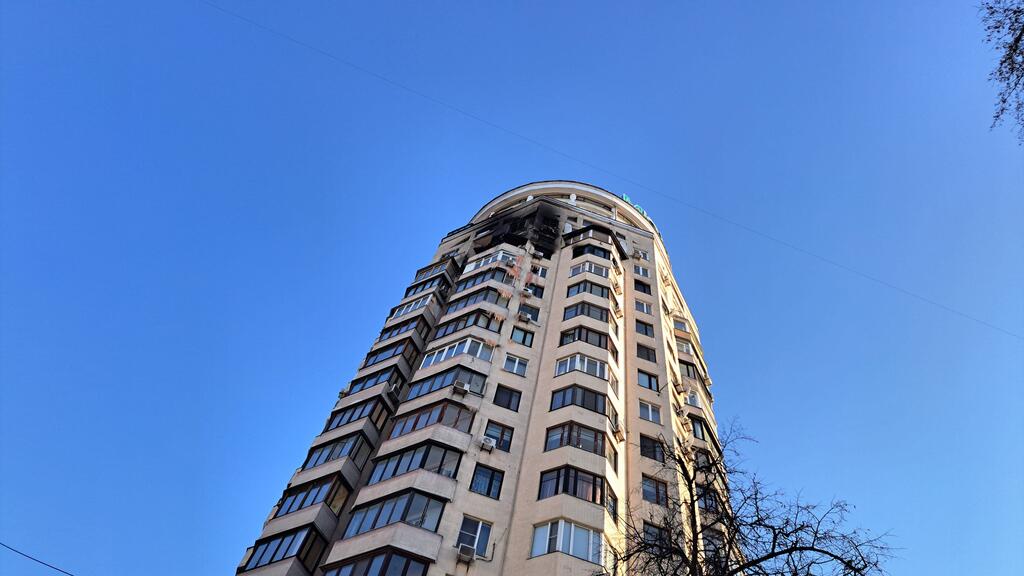 Un haut bâtiment résidentiel en Ukraine avec des dommages visibles à ses étages supérieurs, y compris des fenêtres brisées et des sections charbonnées, contre un ciel bleu clair.