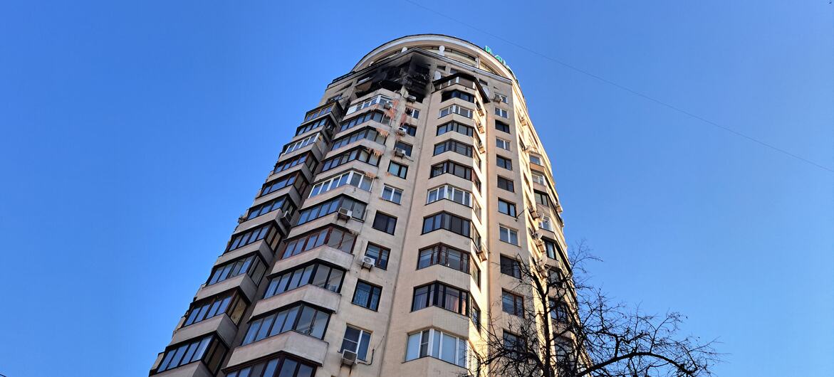 A tall residential building in Ukraine with visible damage to its upper floors, including broken windows and charred sections, against a clear blue sky.