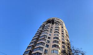 A tall residential building in Ukraine with visible damage to its upper floors, including broken windows and charred sections, against a clear blue sky.