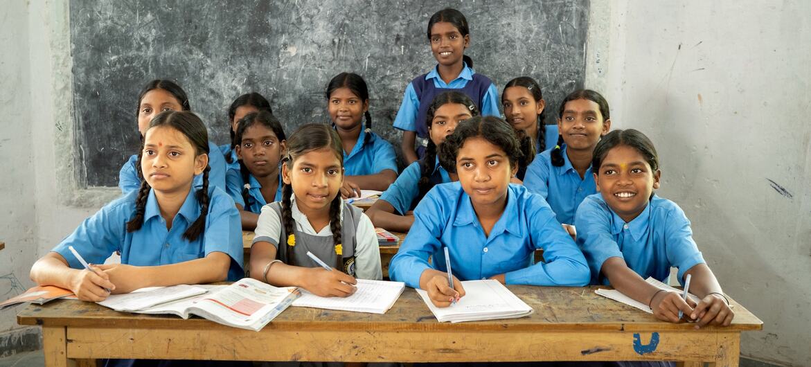 Un groupe de jeunes filles en uniforme scolaire assises à des bureaux dans une salle de classe, avec une fille en tunique grise. L'image capture une initiative éducative inclusive à Purnia, en Inde, soutenue par l'UNICEF et axée sur l'enseignement de la langue des signes indienne pour promouvoir la connexion et la confiance parmi les élèves ayant une déficience auditive.