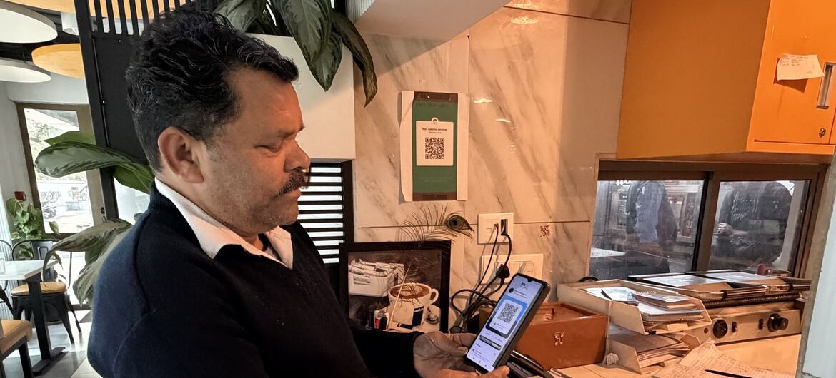 डार्क स्वेटर में एक आदमी पेटीएम क्यूआर कोड स्टैंड के साथ कैफे काउंटर पर अपने स्मार्टफोन पर यूपीआई का उपयोग करके डिजिटल भुगतान करता है।