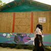 Uma mulher rohingya passa por um vibrante mural em um edifício de bambu em Cox's Bazar, com um sinal "Welcome to Women's Market", simbolizando a resiliência e o empoderamento da comunidade.