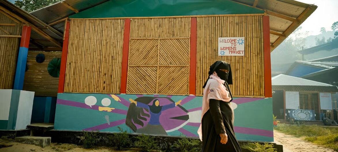 एक रोहिंग्या महिला कोक्स बाजार में एक बांस की इमारत पर एक जीवंत भित्ति चित्र के पास से गुजरती है, जिसमें 'महिला बाजार में आपका स्वागत है' संकेत है, जो समुदाय की लचीलापन और सशक्तिकरण का प्रतीक है।