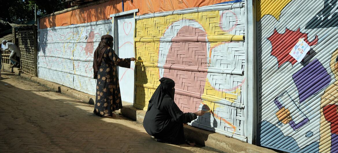 दो रोहिंग्या महिलाएं कॉक्स बाजार में नालीदार धातु की दीवारों पर जीवंत भित्ति चित्र बनाती हैं, कला का उपयोग एकजुटता व्यक्त करने और चल रहे मानवीय संकट के बीच कथनों को बदलने के लिए करती हैं।