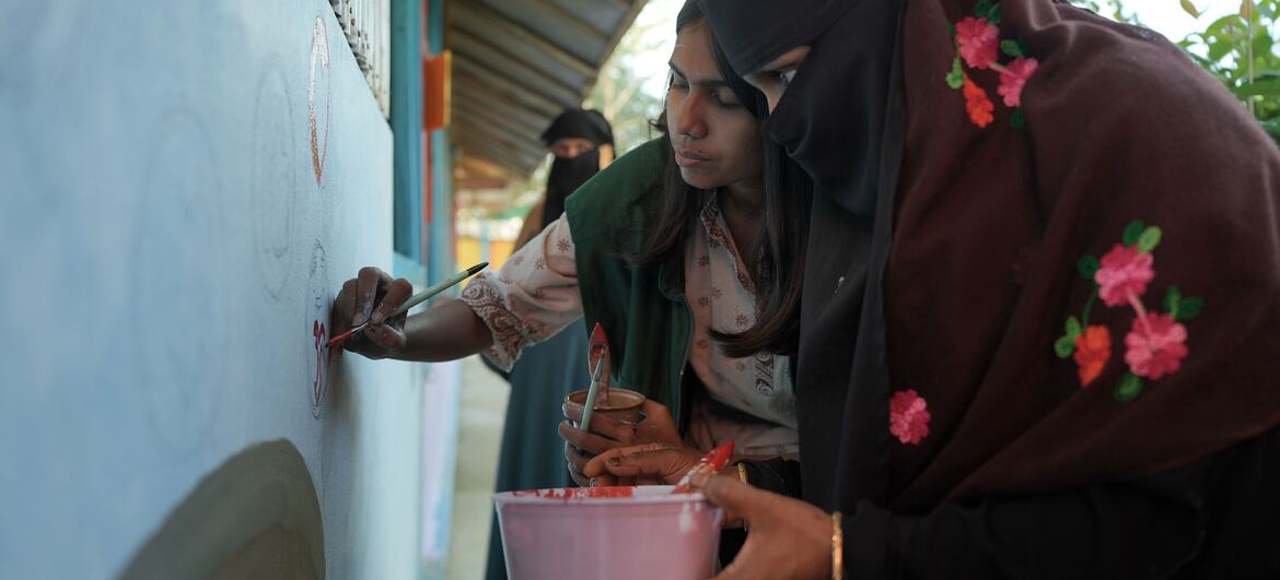 कोक्स बाज़ार में दो रोहिंग्या महिलाएं पेंसिल और पेंट का उपयोग करके दीवार पर एक भित्ति चित्र बना रही हैं, जो एक कहानी कहने की पहल के हिस्से के रूप में एकजुटता बनाने और शरणार्थी संकट के आसपास कथाओं को बदलने के लिए है।