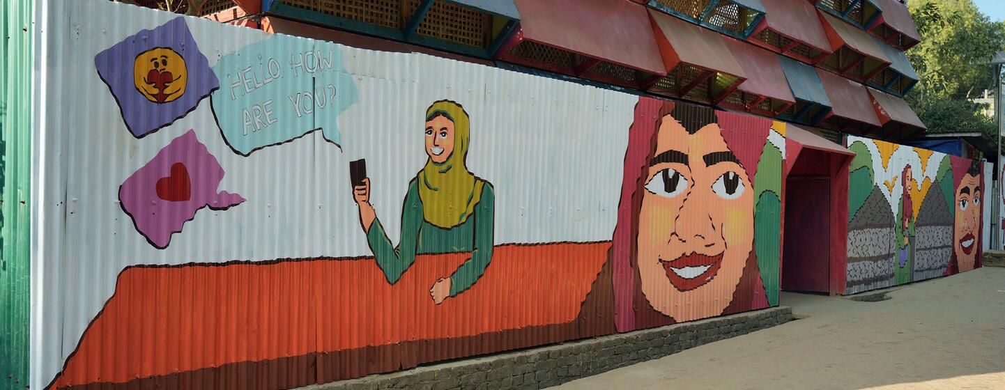 कॉक्स बाजार में एक जीवंत भित्ति चित्र में रोहिंग्या महिलाओं को स्मार्टफ़ोन का उपयोग करते हुए और मुस्कुराते हुए दिखाया गया है, जो चल रहे मानवीय संकट के बीच लचीलापन और एकजुटता का प्रतीक है।