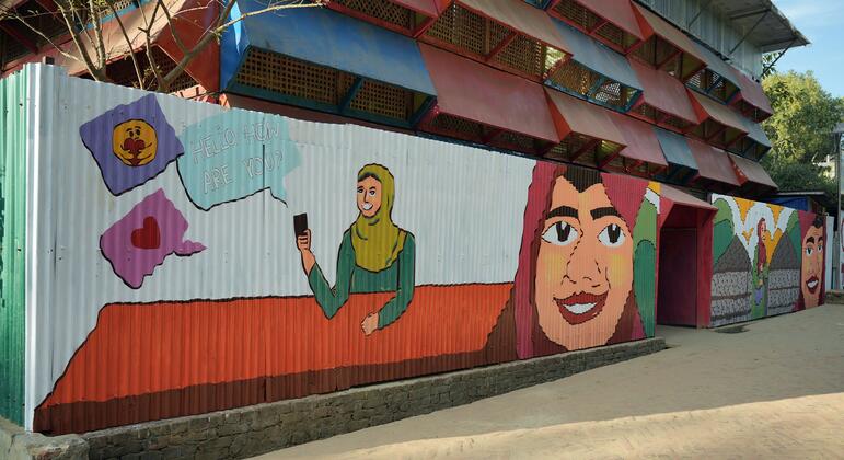 कॉक्स बाजार में एक जीवंत भित्ति चित्र में रोहिंग्या महिलाओं को स्मार्टफ़ोन का उपयोग करते हुए और मुस्कुराते हुए दिखाया गया है, जो चल रहे मानवीय संकट के बीच लचीलापन और एकजुटता का प्रतीक है।