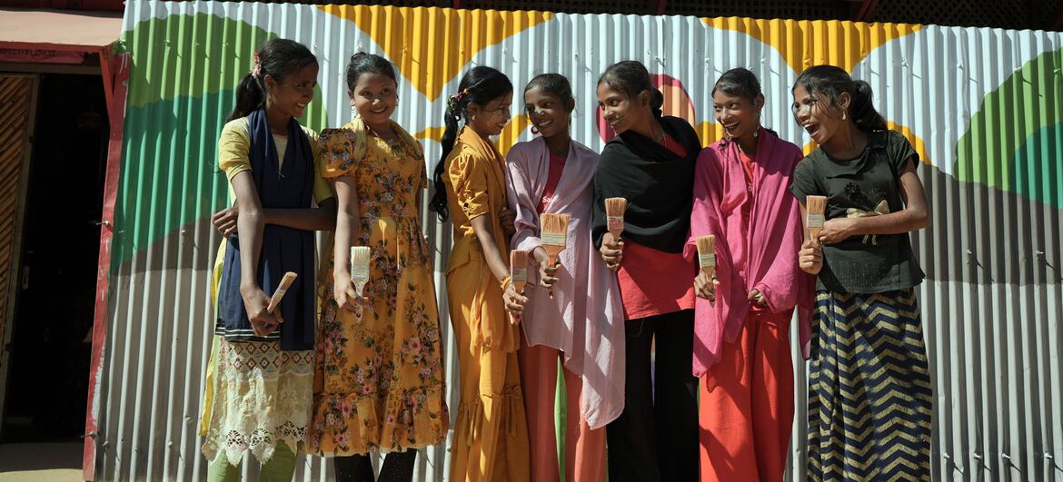 कॉक्स बाज़ार में रोहिंग्या महिलाओं का एक समूह, पेंट ब्रश पकड़ते हुए और मुस्कुराते हुए, कला के माध्यम से अपनी लचीलापन और रचनात्मक अभिव्यक्ति का प्रतीक है।