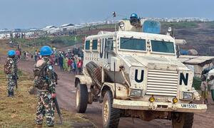A UN peacekeeper in blue helmet and camouflage stands guard next to a large, white UN armored vehicle in Ituri, DRC, with a refugee camp in the background.