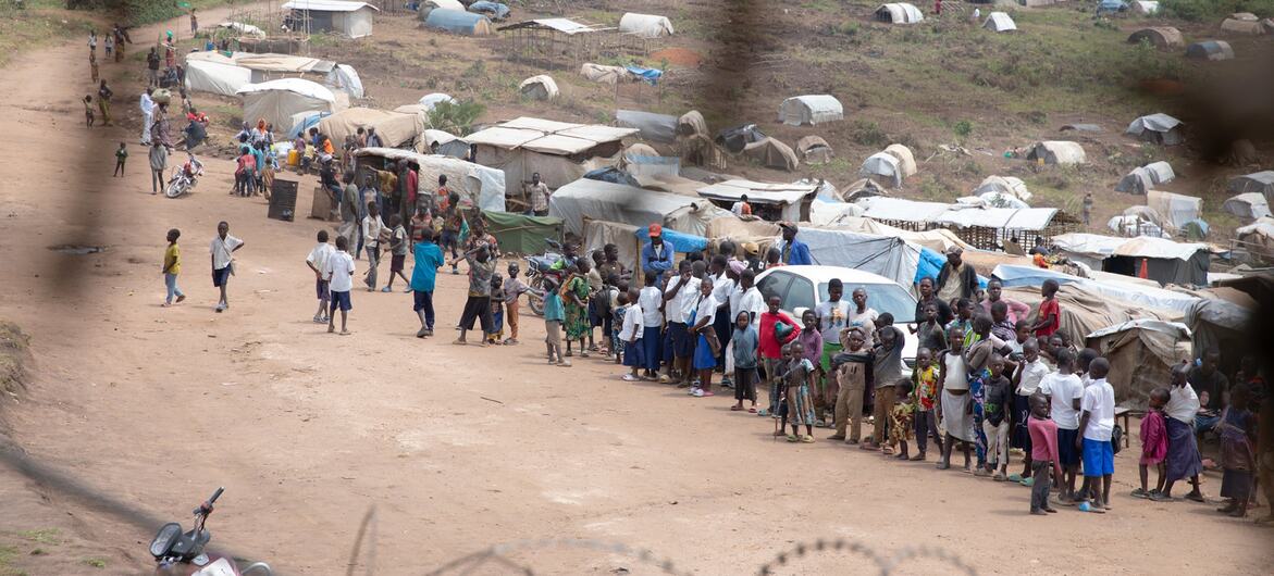 A wide shot of a displacement camp in Ituri, showing numerous temporary shelters and tents on a hillside, with people gathered on a dirt road in the foreground.