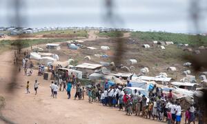 A wide shot of a displacement camp in Ituri, showing numerous temporary shelters and tents on a hillside, with people gathered on a dirt road in the foreground.