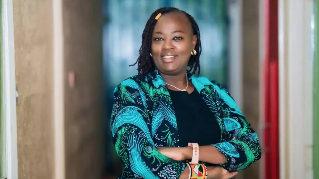 Mwanamke mweusi mwenye tabasamu mwenye nywele zilizotanikwa, amevaa koti lenye rangi ya bluu na kijani kibichi lenye rangi nyangavu juu ya kitambaa nyeusi, amesimama kwenye ukumbi wa maonyesho akiwa na mikono iliyokutanika.