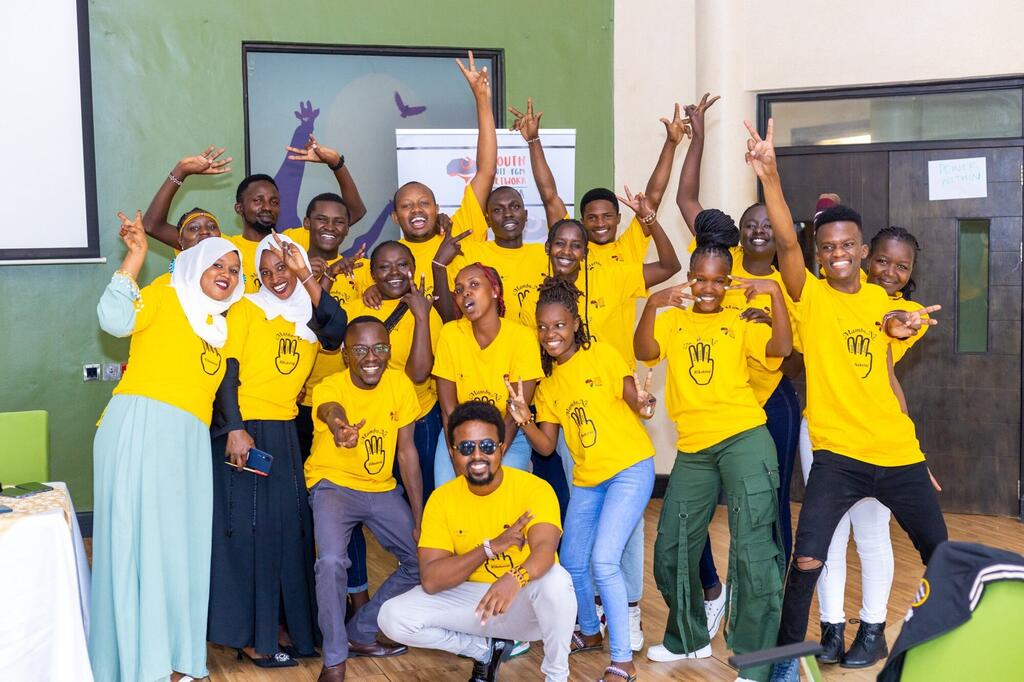 Kikundi cha vijana wenye tabasamu wakiwa wamevaa t-shirts za rangi ya manjano zenye nembo za alama za vidole wakipiga picha kwa shauku kwenye hafla ya kampeni ya UNFPA Kenya ya kukomesha ukandamizaji wa uzazi wa kike.