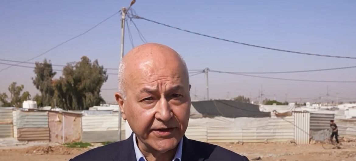A man in a suit speaks to the camera in front of the Zaatari Refugee Camp in Jordan, with temporary shelters and dry landscape in the background.
