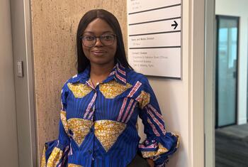 Picha ya mwanaharakati wa haki za binadamu wa Nigeria akiwa amesimama katika ukumbi wa ofisi karibu na ishara ya Idara ya Habari na Vyombo vya Habari ya Umoja wa Mataifa, na nembo ya Umoja wa Mataifa ikionekana ukutani.