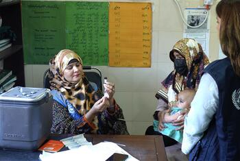 يتحدث عامل صحي تابع لمنظمة الصحة العالمية مع أم تحمل طفلها، بينما يُعدّ مُلقِّح آخر لقاحًا على طاولة مجهزة بالإمدادات الطبية.