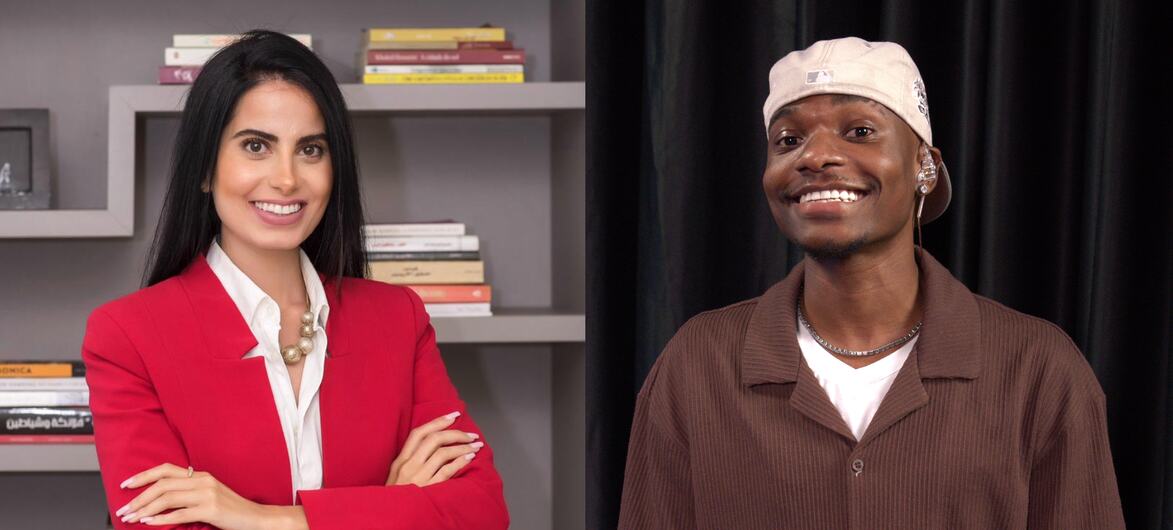 Uma mulher de blazer vermelho e um homem de camisa marrom e boné branco estão sorrindo em um ambiente de estúdio, com estantes de livros ao fundo.