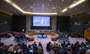 Wide view of the United Nations Security Council meeting discussing threats to international peace and security, with delegates seated around a large oval table and a screen displaying participants via video conference.