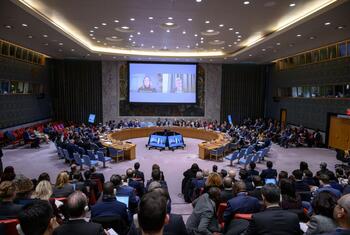 Wide view of the United Nations Security Council meeting discussing threats to international peace and security, with delegates seated around a large oval table and a screen displaying participants via video conference.