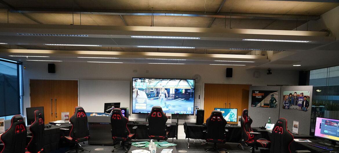 A gaming room at the University of Sussex, featuring multiple gaming stations with black and red chairs, a central table with green pamphlets, and a large screen displaying a video game.