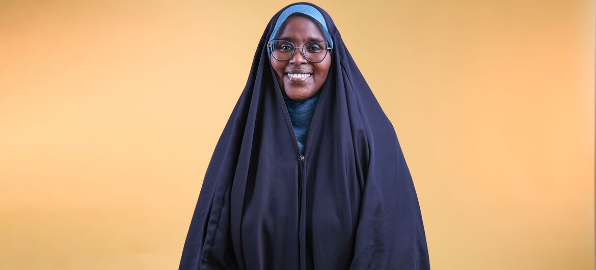 A smiling woman wearing a blue headscarf and glasses, standing against a warm yellow background, representing survivors leading the fight against female genital mutilation in Djibouti.