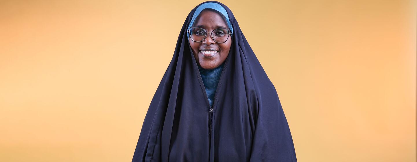 A smiling woman wearing a blue headscarf and glasses, standing against a warm yellow background, representing survivors leading the fight against female genital mutilation in Djibouti.