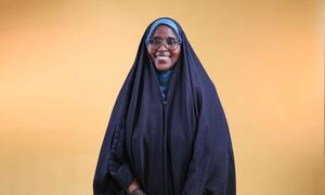 A smiling woman wearing a blue headscarf and glasses, standing against a warm yellow background, representing survivors leading the fight against female genital mutilation in Djibouti.