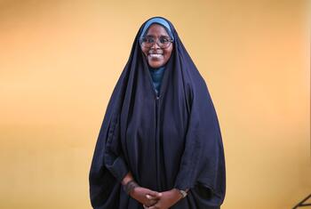 Una mujer sonriente con un pañuelo azul y gafas, de pie sobre un cálido fondo amarillo, representando a las sobrevivientes que lideran la lucha contra la mutilación genital femenina en Djibouti.