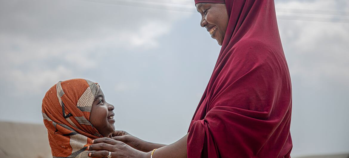 A woman in a red headscarf gently touches the head of a young girl wearing an orange headscarf, both smiling as they look at each other. The image symbolizes resilience and community resistance against female genital mutilation in Somalia.