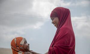A woman in a red headscarf gently touches the head of a young girl wearing an orange headscarf, both smiling as they look at each other. The image symbolizes resilience and community resistance against female genital mutilation in Somalia.
