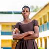 Une jeune femme se tient confiante avec les bras croisés devant un bâtiment scolaire, symbolisant la résilience et la résistance contre la mutilation génitale féminine en Tanzanie.