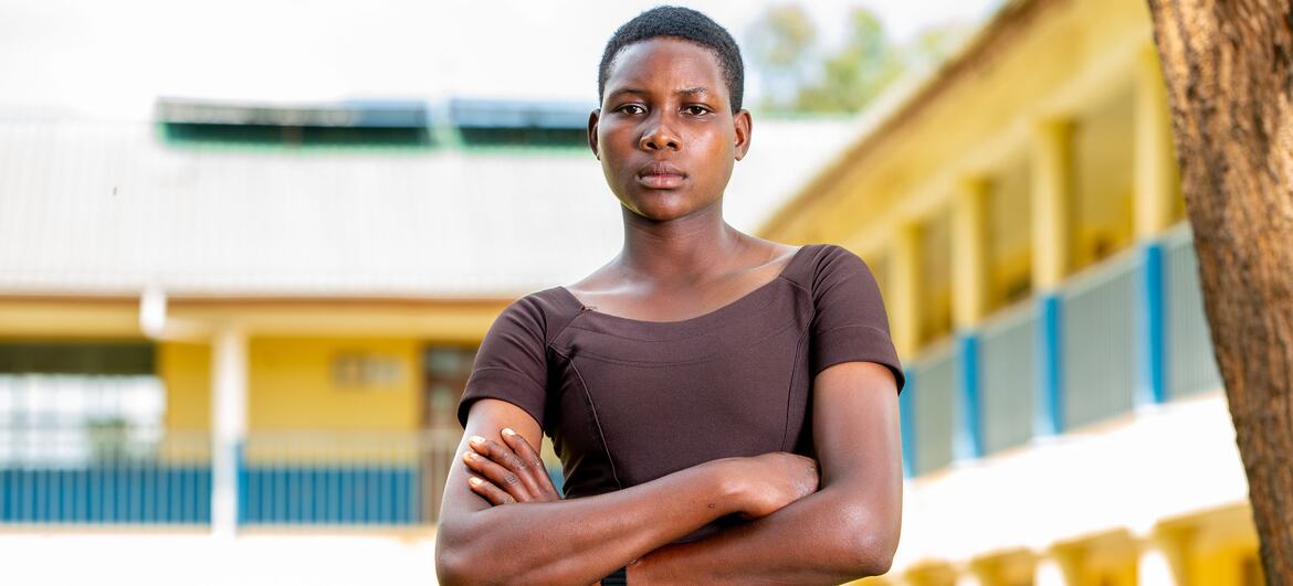 A young woman stands confidently with arms crossed in front of a school building, symbolizing resilience and resistance against female genital mutilation in Tanzania.