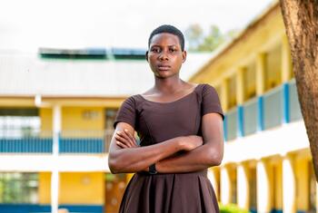 Une jeune femme se tient confiante avec les bras croisés devant un bâtiment scolaire, symbolisant la résilience et la résistance contre la mutilation génitale féminine en Tanzanie.