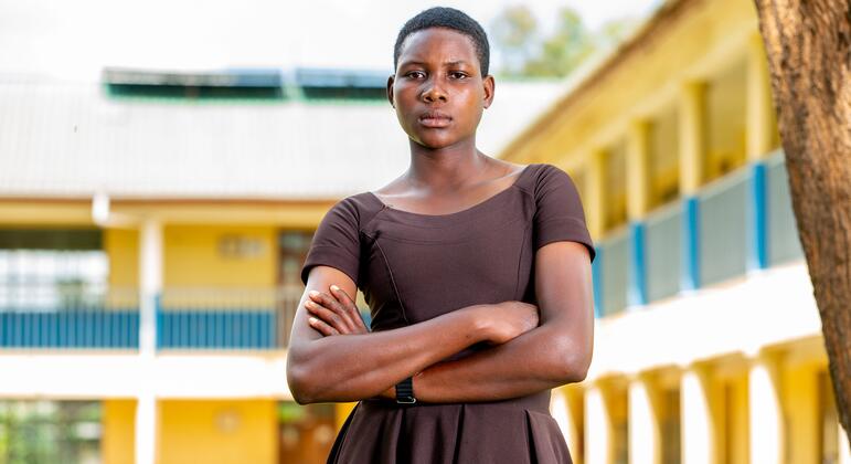 Une jeune femme se tient confiante avec les bras croisés devant un bâtiment scolaire, symbolisant la résilience et la résistance contre la mutilation génitale féminine en Tanzanie.