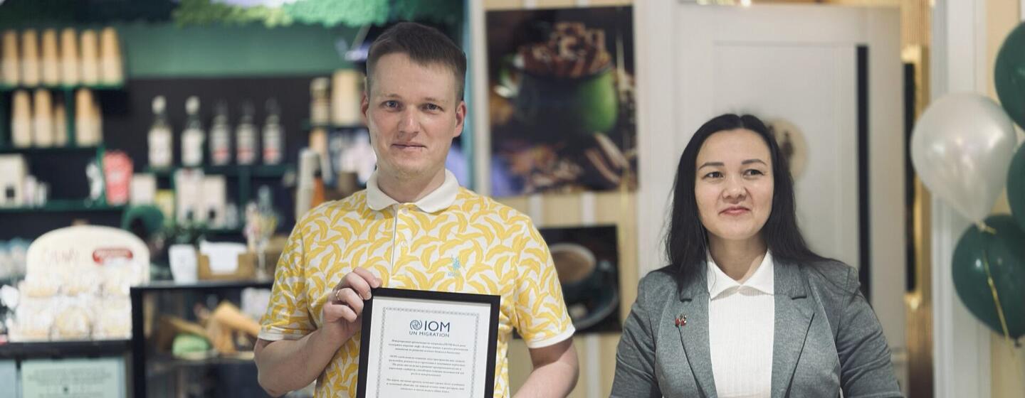 Мужчина в желтой рубашке с рисунком держит в руках каркасный сертификат МОМ (Международная организация по миграции), стоявший рядом с женщиной в сером блейзере.