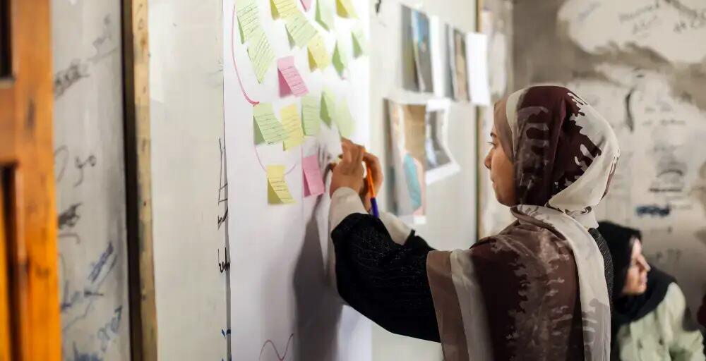 امرأة ترتدي حجاباً على رأسها تشارك بنشاط في جلسة تخطيط، وتكتب على لوحة بيضاء مغطاة بالملصقات الملونة.